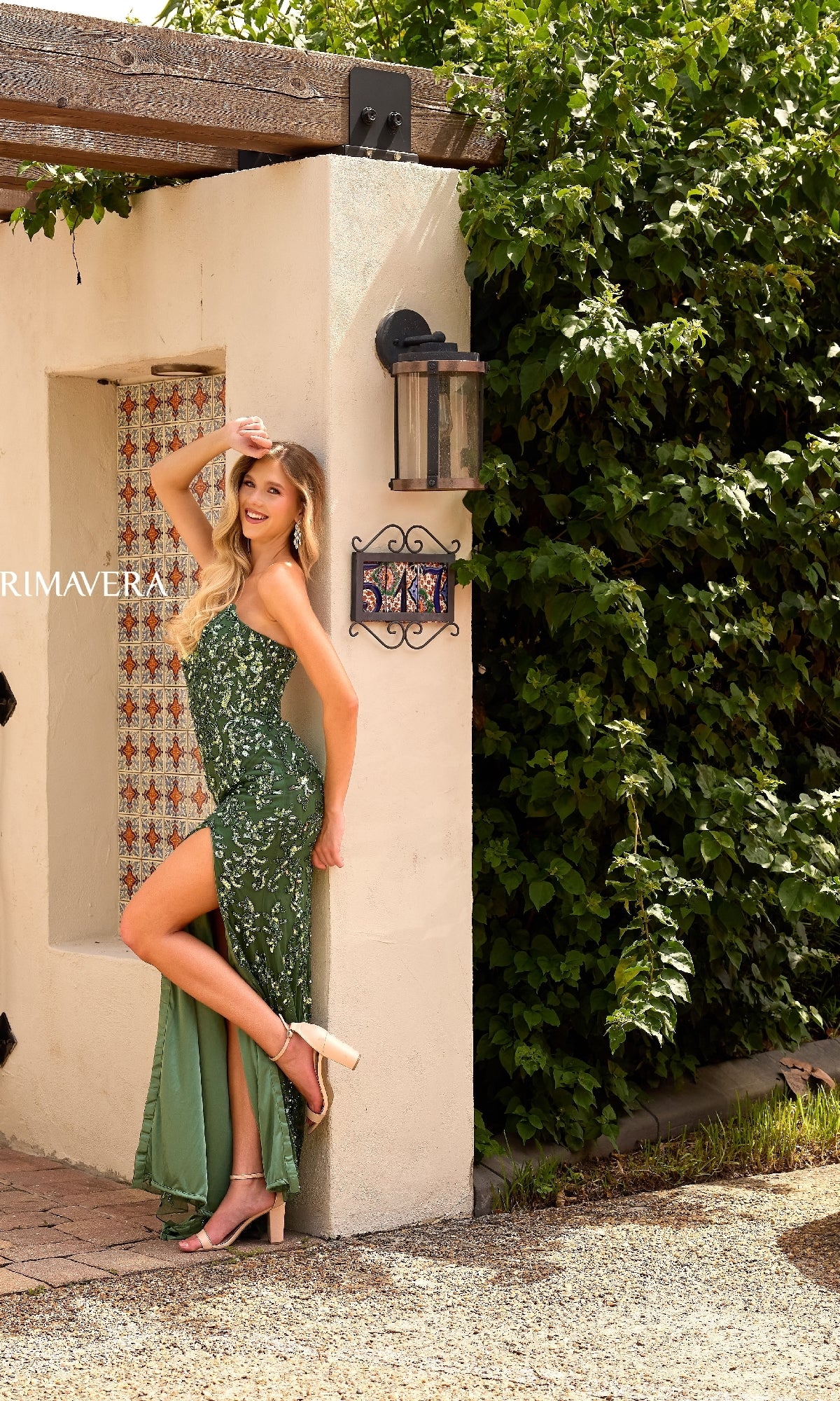 A woman models the Primavera 4347 strapless long sequin prom dress with a thigh-high slit, posing in high-heeled sandals against a stucco wall near greenery.